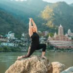 A woman practicing yoga on rocks by the river in Rishikesh, India, with scenic mountain views.