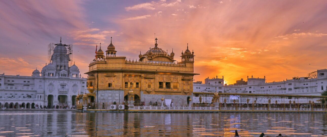 Exterior of Sikh gurdwara golden temple with dome located near water against cloudy sky in evening time in city in India