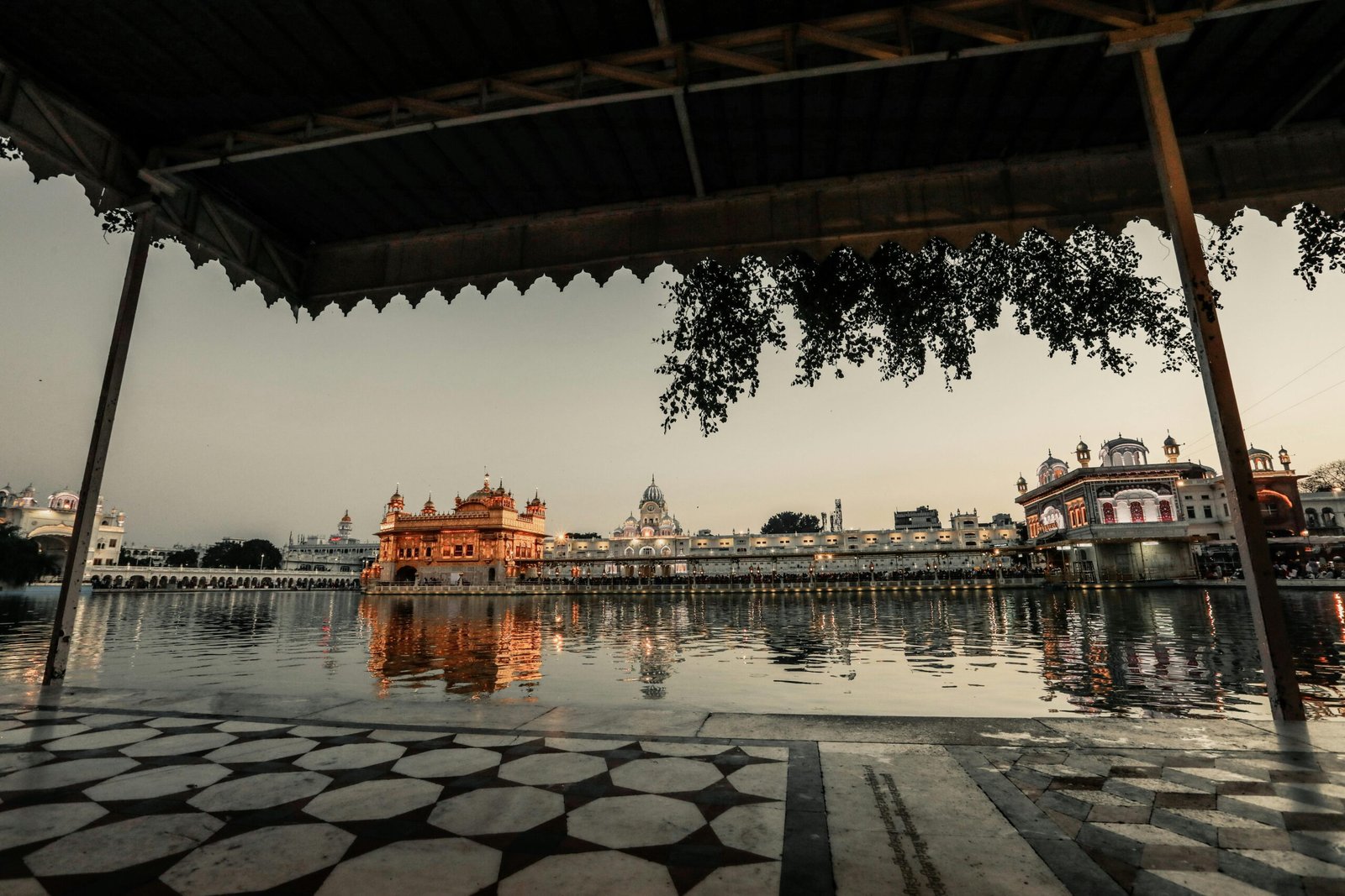 Sidewalk with roof near water with illuminated ancient buildings with fence against gray sky in city street in evening time