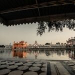 Sidewalk with roof near water with illuminated ancient buildings with fence against gray sky in city street in evening time